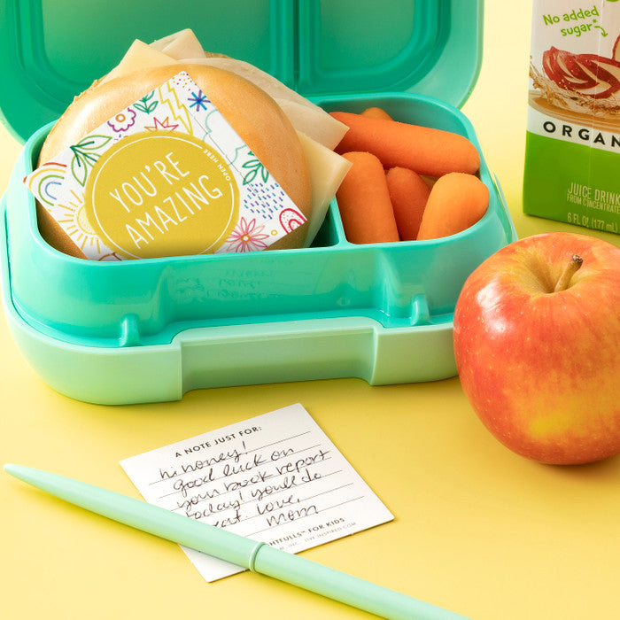 Image of teal lunchbox with thoughtfull card inside and another card with a handwritten note on the table , apples , carrots and a juice box also seen in picture